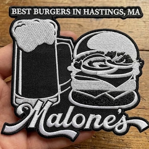 May include: Black and white embroidered patch with a burger and beer mug design. The patch includes the text "BEST BURGERS IN HASTINGS, MA" and "Malone's". The burger has a sesame seed bun and visible fillings, perfect for a Hastings local.