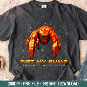 Op de afbeelding: Donkergrijze t-shirt met een opvallend oranje krabontwerp, de tekst "FIST MY BUMP" en "PROJECT HAIL MARY" in goud. De krab wordt omringd door kleine sterren. Het shirt is van het merk Comfort Colors.
