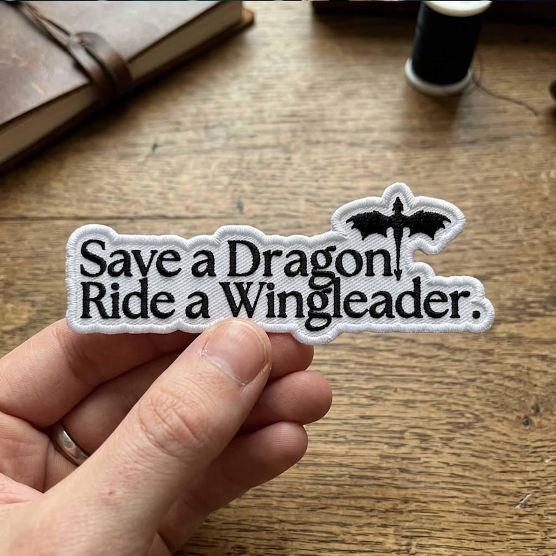 May include: A white embroidered patch with black text that reads "Save a Dragon Ride a Wingleader." The patch features a black dragon silhouette at the top. The patch is held by a hand.