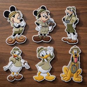 May include: Six embroidered patches featuring cartoon characters in safari outfits. Characters include Mickey Mouse, Minnie Mouse, Goofy, Donald Duck, Daisy Duck, and Pluto. The patches are in shades of brown, tan, and white, with black outlines.