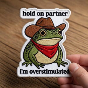 May include: Embroidered patch featuring a cartoon frog wearing a cowboy hat and red bandana. The patch has the text "hold on partner" above the frog and "i'm overstimulated" below. The frog is green with black spots.