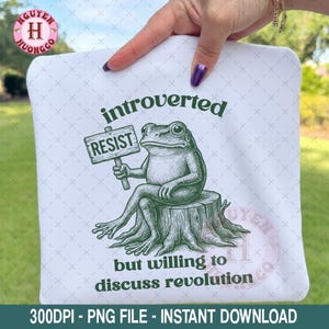 May include: White t-shirt with a green illustration of a frog sitting on a tree stump, holding a sign that says "RESIST." The text above reads "introverted," and below, "but willing to discuss revolution."