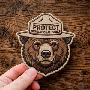 May include: Embroidered patch depicting a brown bear wearing a ranger hat. The hat displays the text "PROTECT OUR NATIONAL PARKS". The bear has detailed brown and tan fur. The patch is held by a hand against a wooden background.