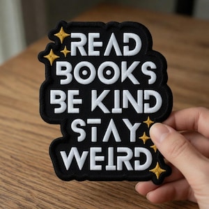 Könnte beinhalten: Schwarzer, gestickter Aufnäher mit den Worten "READ BOOKS BE KIND STAY WEIRD" in weißer Schrift. Der Aufnäher hat einen schwarzen Rand und goldene Sternakzente. Der Aufnäher wird von einer Hand gehalten.