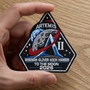 Può includere: Una toppa nera della missione Artemis II. Presenta una luna stilizzata, la Terra e la scritta "ARTEMIS". È visibile anche la scritta "WISEMAN-GLOVER-KOCH-HANSEN TO THE MOON 2026".