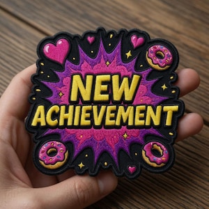 Może przedstawiać: Haftowana naszywka z napisem "NEW ACHIEVEMENT" w kolorze żółtym, na tle fioletowo-różowego wzoru. Naszywka zawiera różowe serca i pączki z posypką. Naszywka jest czarna na krawędziach.