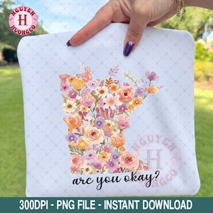 Pode incluir: Um design quadrado azul claro com um arranjo floral em forma do estado de Minnesota. As flores são em tons de rosa, laranja e amarelo. O texto "are you okay?" está abaixo.