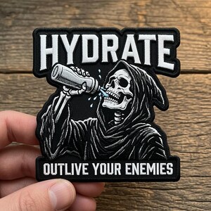 Könnte beinhalten: Ein schwarz-weiß bestickter Aufnäher mit einem Skelett in einer Kapuzenrobe, das aus einer Wasserflasche trinkt. Der Text HYDRATE steht oben und OUTLIVE YOUR ENEMIES unten in weißen Großbuchstaben auf schwarzem Hintergrund.