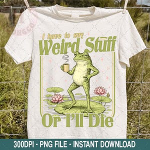 May include: White t-shirt featuring a whimsical illustration of a frog holding a mug, standing on a lily pad. The text reads "I have to say Weird Stuff Or I'll Die" in green. A fun, quirky design for casual wear.