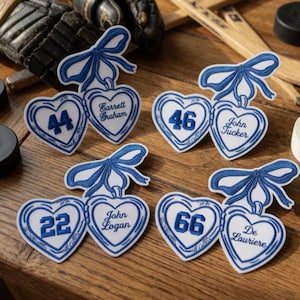 May include: Heart-shaped patches in white and blue, each with a number and a name, topped with a blue bow. The patches have a stitched edge and include names such as "John Tucker" and numbers like "46".