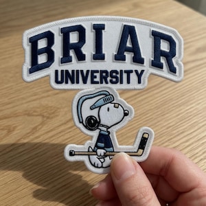 May include: Embroidered patch featuring the text "BRIAR UNIVERSITY" in navy blue on a white background. Below the text is a cartoon character wearing a hockey helmet and holding a hockey stick. The patch has a white border.