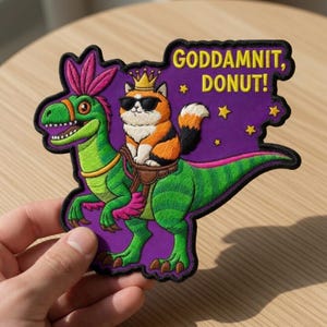 May include: Embroidered patch featuring a cat wearing sunglasses and a crown riding a green dinosaur. The patch has the text "GODDAMNIT, DONUT!" in yellow. The background is purple with yellow stars, and the patch has a black border.