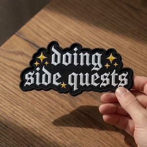 Puede incluir: Parche bordado negro con las palabras "doing side quests" en letras góticas blancas. El parche tiene un borde negro con detalles de estrellas amarillas. El parche está sostenido por una mano y descansa sobre una superficie de madera.