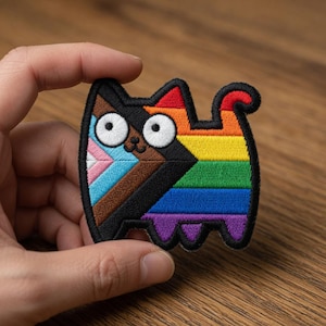 May include: Embroidered cat patch featuring a rainbow design with the Progress Pride flag colors. The cat has large white eyes and a black outline. The patch is held by a hand.