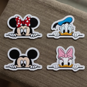 Puede incluir: Cuatro parches bordados con personajes de Disney: Minnie Mouse con un lazo rojo de lunares, el Pato Donald con un sombrero azul, Mickey Mouse y Daisy con un lazo rosa. Cada personaje asoma por encima de un borde blanco.