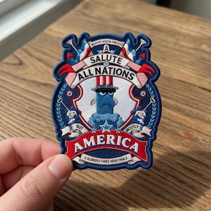 Può includere: Toppa ricamata con un design blu, rosso e bianco. La toppa presenta il testo "A Salute to All Nations, But Mostly America". Un personaggio blu con un cappello a cilindro è al centro. La toppa ha la forma di uno scudo.