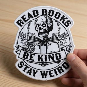 Könnte beinhalten: Ein weißer Aufnäher mit schwarzem Text und einem Totenkopf, der ein Buch liest. Der Aufnäher trägt die Aufschrift "READ BOOKS, BE KIND, STAY WEIRD" in einem kreisförmigen Design mit einem Banner. Der Totenkopf ist von Blättern umgeben.