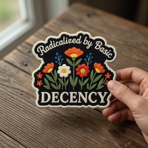 Puede incluir: Parche bordado con fondo negro y diseño floral. El parche presenta flores naranjas, rojas, blancas y azules con hojas verdes. El texto "Radicalized by Basic" está arqueado en la parte superior y "DECENCY" en la inferior.