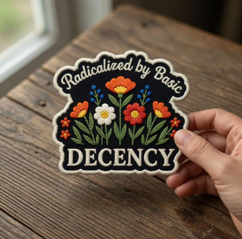 K&ouml;nnte beinhalten: Gestickter Aufn&auml;her mit schwarzem Hintergrund und floralem Design. Der Aufn&auml;her zeigt orange, rote, wei&szlig;e und blaue Blumen mit gr&uuml;nen Bl&auml;ttern. Der Text "Radicalized by Basic" ist oben gew&ouml;lbt und "DECENCY" unten.