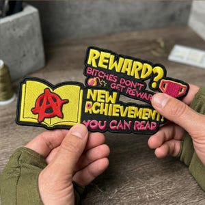 Könnte beinhalten: Zwei gestickte Aufnäher mit schwarzen Rändern. Einer zeigt ein gelbes Buch mit einem roten Anarchie-Symbol. Der andere hat den Text "REWARD?" und "NEW ACHIEVEMENT: YOU CAN READ" in Gelb und Pink.