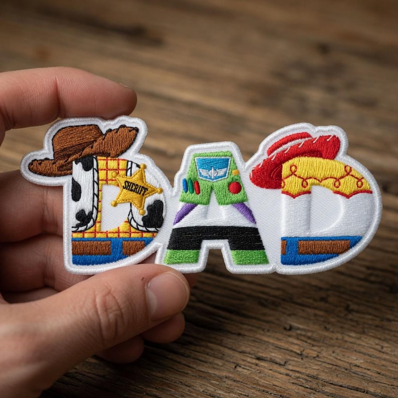 May include: Embroidered patch spelling "DAD" with letters designed as characters from the Toy Story movie. The "D" is Woody, the "A" is Buzz Lightyear, and the second "D" is Jessie. The patch is white with colorful accents.