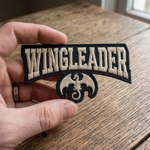 Pode incluir: Patch bordado preto e bege com a palavra "WINGLEADER" em letras bege. Abaixo, um emblema de dragão bege. O patch é mostrado em um fundo de madeira.