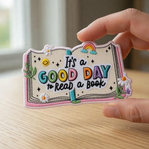 Könnte beinhalten: Gestickter Aufnäher in Form eines offenen Buches mit dem Text "It's a GOOD DAY to READ a BOOK". Der Aufnäher zeigt einen Regenbogen, Gänseblümchen und eine lächelnde Sonne. Der Rand ist rosa.