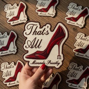Puede incluir: Parches bordados con un diseño de zapato de tacón alto burdeos, el texto "That's All" y "Miranda Priestly". Los parches tienen un fondo color crema y un borde marrón. Los parches están dispuestos sobre una superficie de madera.
