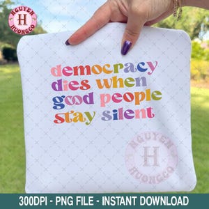 May include: White t-shirt featuring the phrase "democracy dies when good people stay silent" in various colours. The shirt is held by a person with a purple nail. The image also includes a logo and text at the bottom.