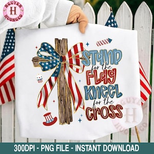 May include: White graphic tee featuring a wooden cross adorned with a red, white, and blue ribbon. The text reads "Stand for the Flag, Kneel for the Cross." Patriotic design with fireworks and a hat.