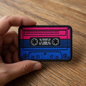 May include: A rectangular embroidered patch in the shape of a cassette tape. The top section is pink, the middle is blue, and the bottom is blue. The cassette tape has a black border and gray details.