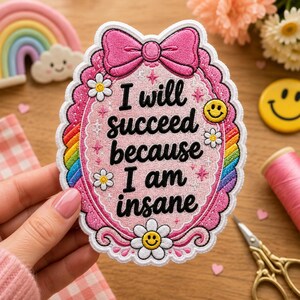 Può includere: Una toppa ricamata ovale con un bordo smerlato rosa e un grande fiocco rosa in alto. Il centro è rosa chiaro con il testo nero "I will succeed because I am insane" circondato da piccole margherite bianche, stelline e un bordo arcobaleno.