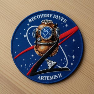 Op de afbeelding: Een cirkelvormige blauwe patch met een vintage duikhelm in bruin- en goudtinten. De tekst "RECOVERY DIVER" is bovenaan gebogen, met "ARTEMIS II" onderaan. Rode en witte accenten maken het ontwerp compleet.