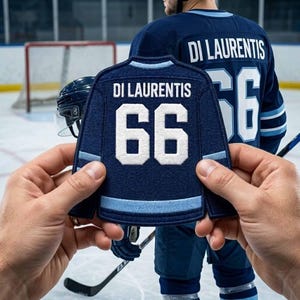Pode incluir: Um patch de camisa de hóquei azul marinho com o nome "DI LAURENTIS" e o número "66" em branco. O patch é segurado em frente a uma pessoa vestindo uma camisa combinando. Um capacete e um taco de hóquei também são visíveis.