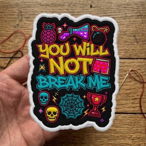 Może przedstawiać: Haftowana naszywka z napisem "YOU WILL NOT BREAK ME" w kolorze żółtym i turkusowym. Naszywka zawiera kolorowe ilustracje ananasa, czaszek, zlewki, bomby i trofeum. Naszywka ma czarne tło z białą obwódką.