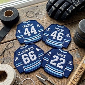 Puede incluir: Cuatro parches de camiseta de hockey azul marino con números y nombres blancos: "GRAHAM 44", "TUCKER 46", "DI LAURENTIS 66" y "LOGAN 22". Equipo de hockey y cinta adhesiva rodean los parches sobre una superficie de madera.