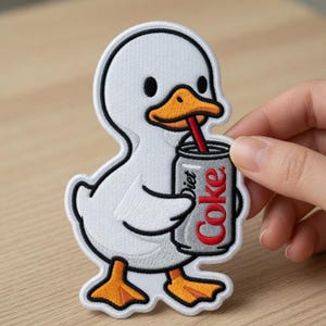 Może przedstawiać: Haftowana naszywka przedstawiająca białą kaczkę z pomarańczowym dziobem i stopami, trzymającą puszkę Diet Coke z czerwoną słomką. Naszywka ma czarny kontur i znajduje się na drewnianej powierzchni.