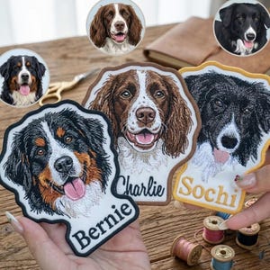 May include: Three embroidered dog patches featuring portraits of a Bernese Mountain Dog, a brown and white spaniel, and a black and white dog. The patches include the names Bernie, Charlie, and Sochi stitched in black or gold thread.