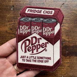 Puede incluir: Parche bordado que representa una caja granate de "Fridge Cigs" con tres latas de refresco "DDr leppe". El parche también muestra el logotipo de "Dr Pepper" y la frase "JUST A LITTLE SOMETHING TO TAKE THE EDGE OFF."