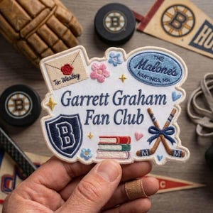 May include: Embroidered patch with the text "Garrett Graham Fan Club." The patch features a shield with a "B," crossed hockey sticks, books, and floral accents. The patch also includes the text "The Malone's Hastings, MA."