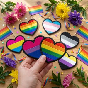 Pode incluir: Uma coleção de emblemas de tecido em forma de coração com vários designs de bandeiras do orgulho, incluindo padrões arco-íris, transgénero, pansexual, bissexual, assexual e não binário, dispostos com pequenas bandeiras arco-íris e flores.