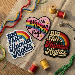 May include: Three embroidered patches with the text "BIG FAN OF Human Rights" and rainbow designs. One patch is heart-shaped, another is a rounded rectangle, and the third is a semi-circle. Spools of thread and sewing tools are scattered around the patches.