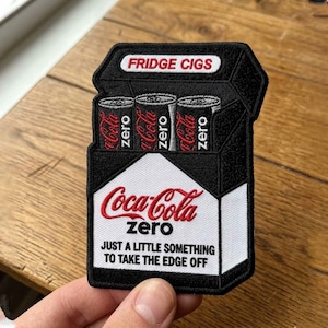 Puede incluir: Parche bordado que representa un paquete de cigarrillos negro con tres latas de Coca-Cola Zero. El parche dice "FRIDGE CIGS" en la parte superior y "Coca-Cola Zero" con la frase "JUST A LITTLE SOMETHING TO TAKE THE EDGE OFF" debajo.