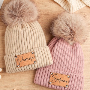 Puede incluir: Dos gorros de punto con pompones de piel sintética. Uno es beige, el otro rosa. Cada gorro tiene un parche de cuero con un nombre. El gorro beige dice "Pamela" y el gorro rosa dice "Salome".