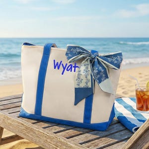 May include: A canvas tote bag with blue trim and handles, personalized with the name "Wyatt" in blue script. A blue and white patterned scarf is tied to the handle. The bag sits on a wooden beach chair near the ocean.