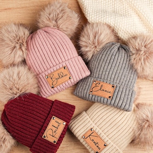 May include: Four ribbed knit beanies in pink, gray, burgundy, and beige, each with a faux fur pom-pom and a rectangular patch with a name. The names are "Baldie", "Elanie", "Rachel", and "Kenton".