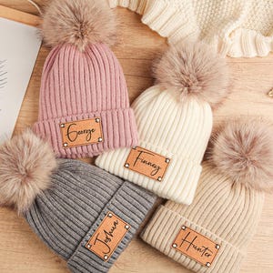 May include: A collection of ribbed knit beanies in pink, cream, gray, and tan, each topped with a fluffy pom-pom. Each beanie has a leather patch with a name on it. A framed print with the words "BE BRAVE" and a lion illustration is in the background.