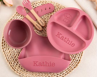 以下が含まれることがあります： ダスティローズ色のシリコン製ベビー食器セット。3つに仕切られたプレート、ボウル、ビブ、木製ハンドルのスプーンとフォークが含まれます。プレートとビブには「Kathie」という名前と子犬のグラフィックが刻印されています。