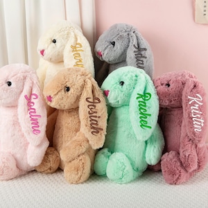 May include: A collection of plush bunny rabbits in various pastel colors, including pink, beige, and mint green. Each bunny has long, floppy ears with a name embroidered on one ear. The bunnies are soft and cuddly.