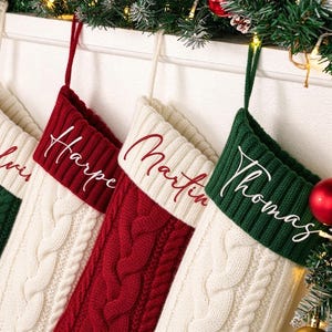 May include: Four cable-knit Christmas stockings hanging in a row. The stockings alternate between green and red bodies with contrasting white or green cuffs. Each cuff features a personalized name in white or red script: Calvin, Harper, Martin, and Thomas.